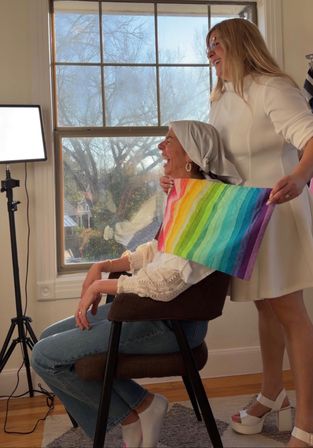 Sunlit indoor portrait of a seated woman wearing a headscarf laughing as a stylist holds a rainbow color drape against her shoulders by a large window, natural light and cheerful color-consultation vibe.