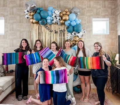 Smiling group of women in a living room posing under a blue, gold and cow‑print balloon arch with gold fringe, each holding colorful striped fabric swatches at an indoor celebration