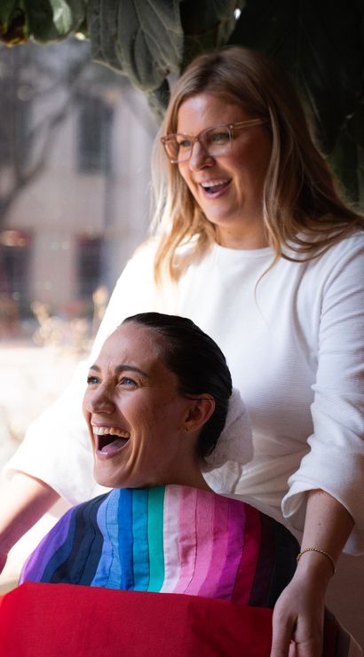 Laughing client wrapped in a colorful salon cape with a smiling stylist standing behind her in a bright, window-lit salon