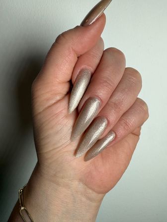Close-up of a hand with long stiletto-shaped nails painted in shimmering champagne-gold polish, wrist bracelet visible against a neutral background.