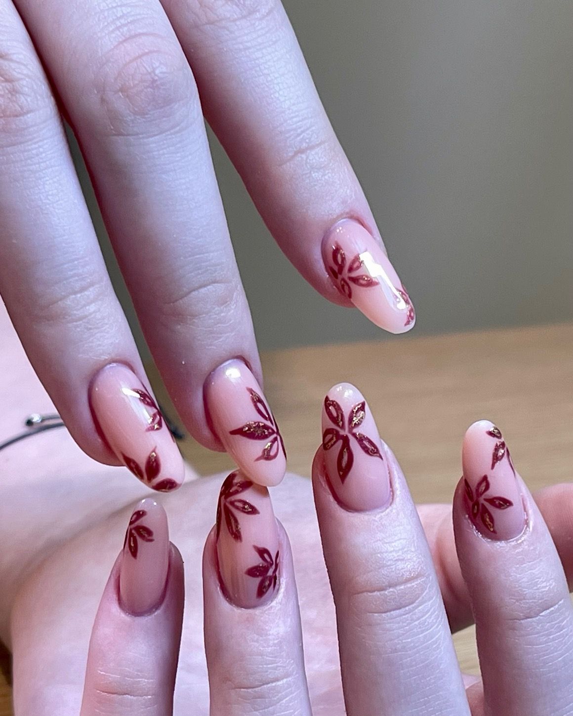 Close-up of hands with almond-shaped nude-pink manicure featuring maroon floral leaf designs and subtle gold accents, modern salon nail art.
