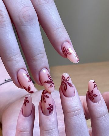 Close-up of hands with almond-shaped nude-pink manicure featuring maroon floral leaf designs and subtle gold accents, modern salon nail art.