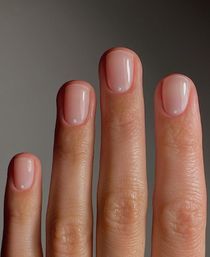 Close-up of four fingers showcasing a minimalist short glossy nude-pink manicure with tiny white dot accents near the cuticles on a neutral gray background.