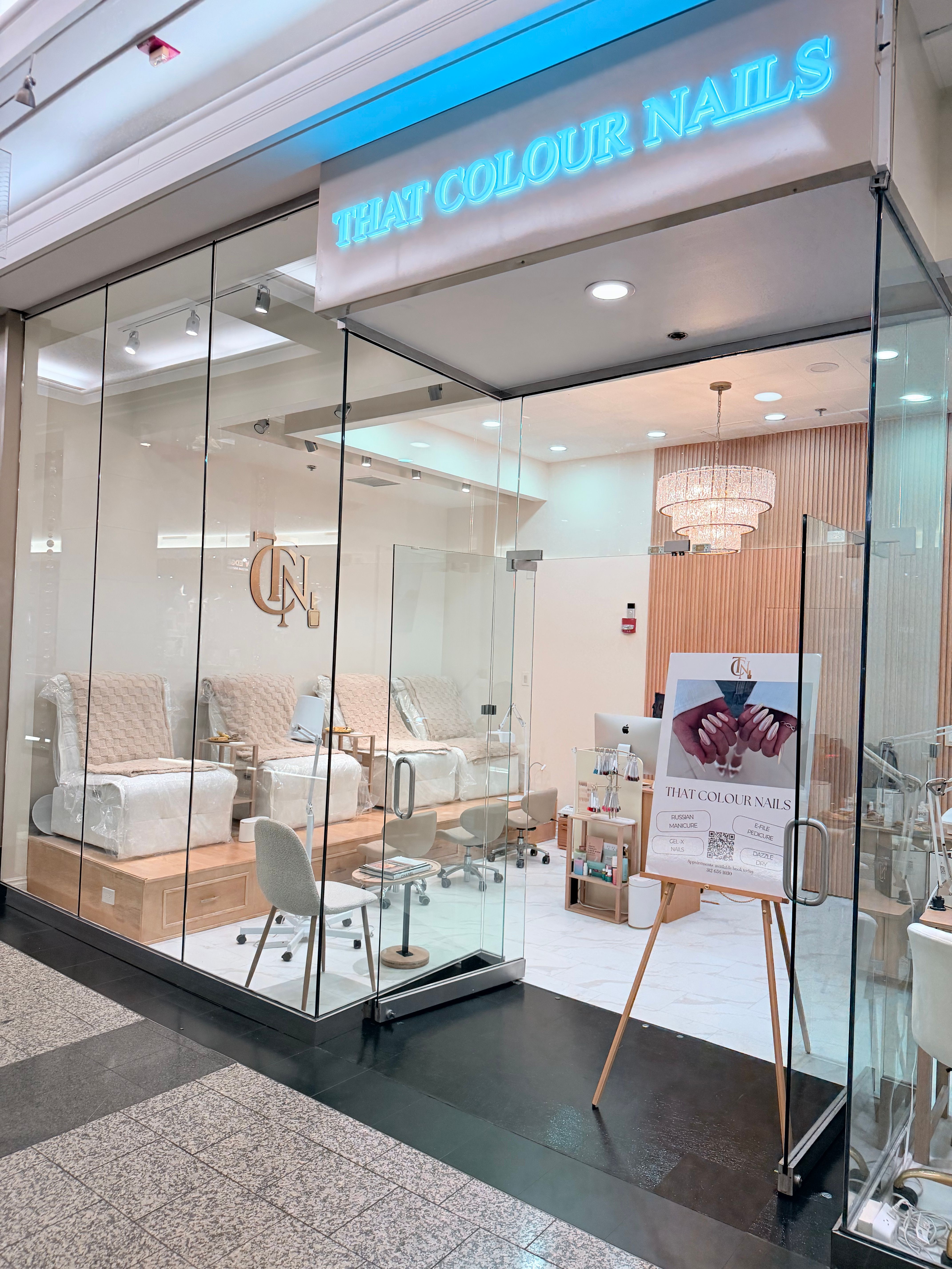Modern mall nail salon behind a glass storefront with plush pedicure chairs on a raised wooden platform, neutral decor, crystal chandelier, and an easel sign showing manicured nails.