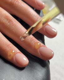 Close-up of manicured fingers with glossy nude nails as golden cuticle oil drops from a dropper onto the cuticles during a manicure.