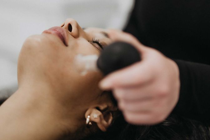 Close-up of a client relaxing during a spa facial while an esthetician applies cream with a round massage tool to the cheek — skincare treatment and pampering.