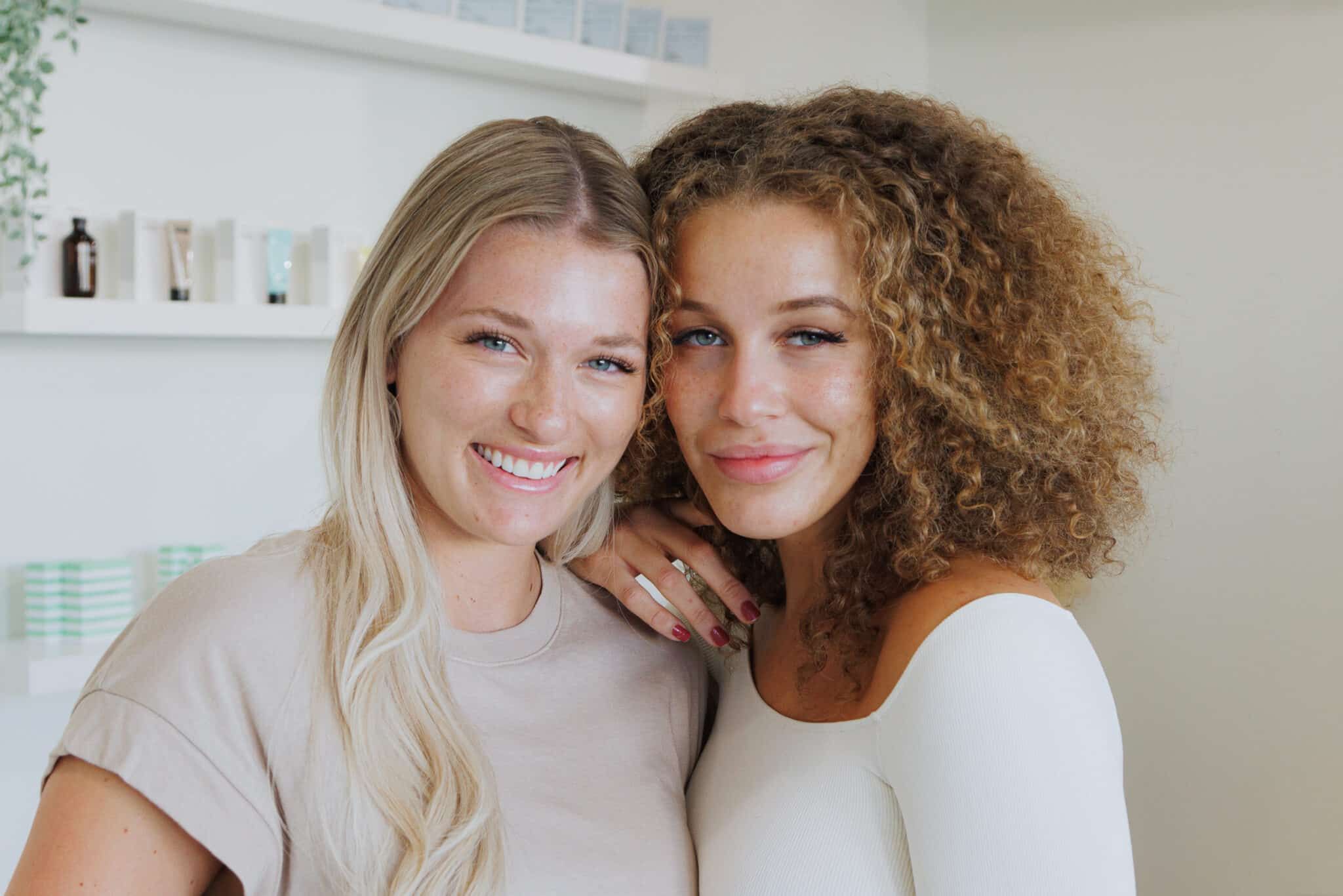 Close-up portrait of two smiling women posing together in a bright beauty studio with shelves of skincare products
