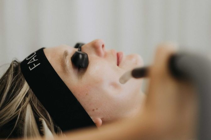 Close-up spa facial: client wearing a black headband and small eye shields receiving a handheld skin treatment on the jawline.