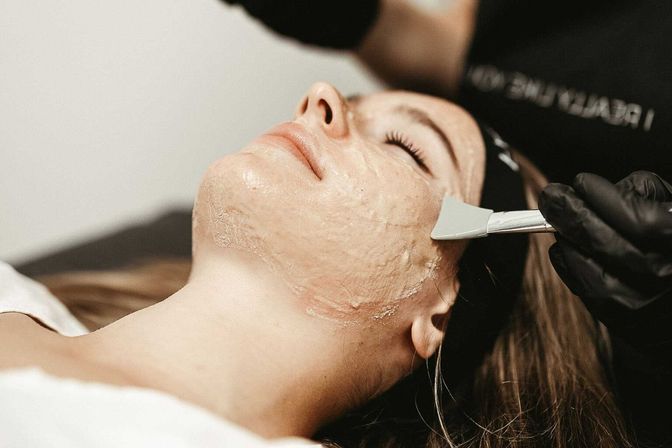 Close-up of a client with eyes closed as an esthetician in black gloves spreads an exfoliating facial mask with a spatula during a relaxing spa facial treatment