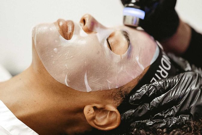 Close-up spa facial: client wearing a translucent hydrogel sheet mask while an esthetician in black gloves uses a handheld LED/microcurrent device for a rejuvenating treatment.