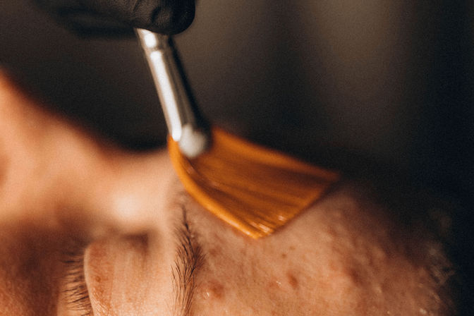Close-up of a gloved hand using a fan brush to apply a golden facial mask to a client's closed eye and cheek during a spa skincare treatment