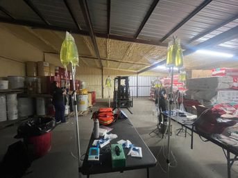 Temporary medical setup in an industrial warehouse with multiple IV drip bags hanging from poles above tables holding medical supplies, emergency backpacks, and equipment amid stacked pallets, barrels, and a forklift.