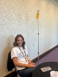 Smiling woman seated in a conference room receiving IV hydration from a yellow infusion bag on a pole, with a smartphone and cup on the table
