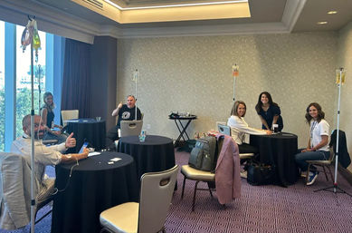 Adults receiving IV infusions at round tables in a bright hotel conference room with city view, smiling and giving thumbs-up
