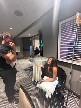 Laughing woman seated in a hotel conference hallway with a pillow and IV drip bag, chatting with a standing man holding a pen and clipboard near a round cocktail table and framed wall art.
