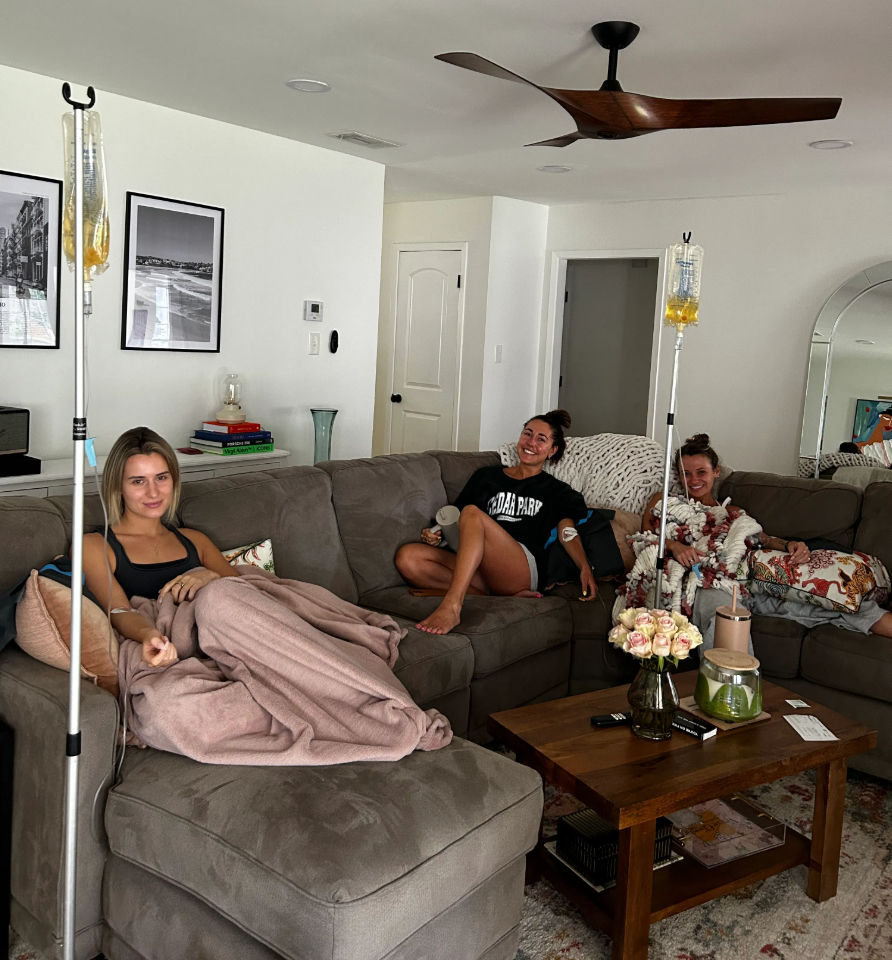 Three friends lounging on a gray sectional in a bright home living room, each receiving at-home IV drips from poles; wooden coffee table with vase of pale roses, books and decor, ceiling fan overhead and cozy blankets and pillows.