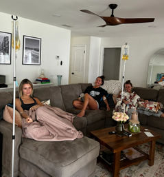 Three friends lounging on a gray sectional in a bright home living room, each receiving at-home IV drips from poles; wooden coffee table with vase of pale roses, books and decor, ceiling fan overhead and cozy blankets and pillows.