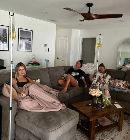 Three friends lounging on a gray sectional in a bright home living room, each receiving at-home IV drips from poles; wooden coffee table with vase of pale roses, books and decor, ceiling fan overhead and cozy blankets and pillows.