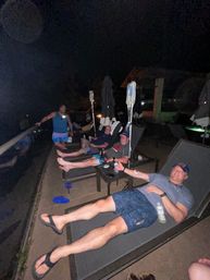 Group of men relaxing on an outdoor pool deck at night on lounge chairs, two IV drip poles nearby, holding drinks.