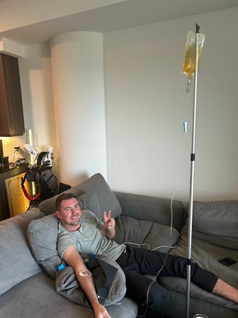 Smiling man reclining on a gray couch in a modern apartment living room receiving at-home IV infusion from a tall pole with a yellow IV bag, flashing a peace sign; golf bag and kitchenette visible in the background.