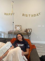 Smiling woman reclining on a living-room couch receiving a home IV infusion, cozy blanket and orange throw, with a glittery "Happy Birthday" banner and a "Bless this Home" sign on the wall.