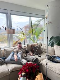 Smiling man relaxing on a beige sectional in a high-rise city apartment receiving IV fluids from a pole, with large windows showing a skyline view, indoor palm plants and a red poinsettia on the coffee table.