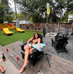 Woman in green bikini reclines in a black Adirondack chair on a backyard patio, smiling while receiving an IV hydration drip from a yellow bag on a pole; outdoor lounge area with artificial turf, bright yellow chaise loungers, striped umbrella, wooden fence, fire pit, and drinks on the deck.