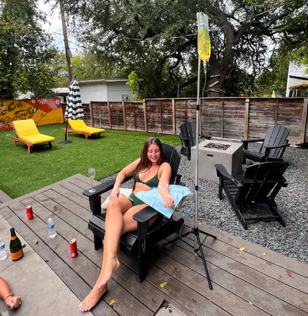 Woman in green bikini reclines in a black Adirondack chair on a backyard patio, smiling while receiving an IV hydration drip from a yellow bag on a pole; outdoor lounge area with artificial turf, bright yellow chaise loungers, striped umbrella, wooden fence, fire pit, and drinks on the deck.
