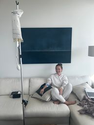 Smiling woman receiving at-home IV infusion while lounging on a white sectional couch, with an IV pole and portable pump beside her, blue abstract wall art behind and a laptop nearby — cozy living room wellness setup.