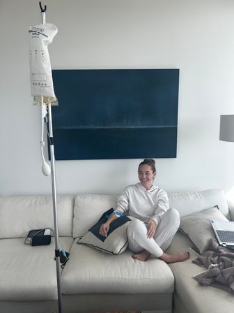 Smiling woman receiving at-home IV infusion while lounging on a white sectional couch, with an IV pole and portable pump beside her, blue abstract wall art behind and a laptop nearby — cozy living room wellness setup.