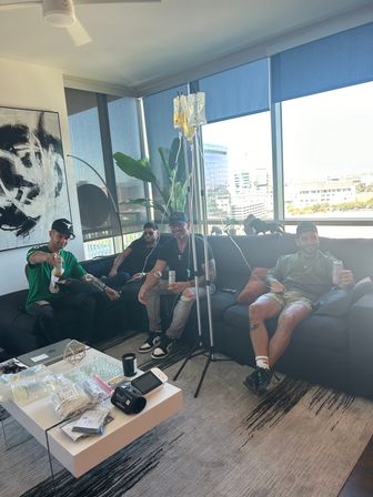 Three people lounging on a high-rise apartment couch receiving IV drips from a pole while holding drinks, medical supplies on a coffee table and a city skyline visible through floor-to-ceiling windows.