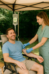 Smiling man reclines under a pop-up canopy while a nurse in green scrubs and gloves administers an IV drip — outdoor mobile IV therapy and wellness treatment.