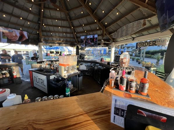 Open-air waterfront tiki-style bar under a wooden pavilion with string lights, wooden counter, drink dispensers and liquor bottles, TVs overhead and outdoor patio seating with blue umbrellas.