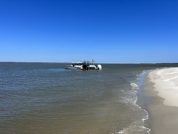 Small motorboat anchored off a sandy beach on a calm coastal bay, gentle waves lapping the shore under a clear blue sky.