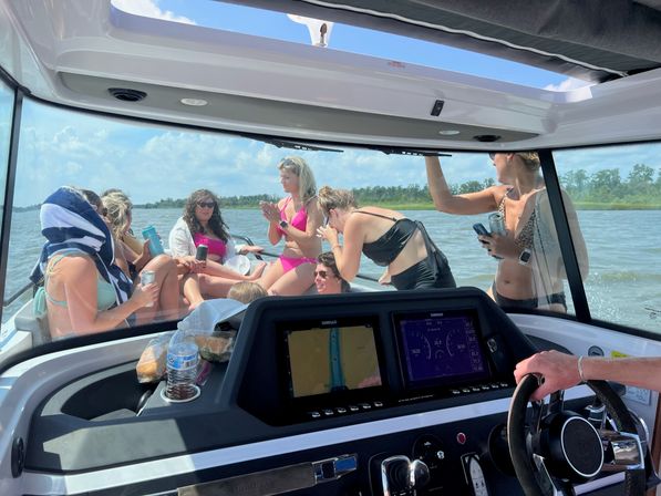 Group of friends in swimsuits lounging at the bow of a motorboat on a sunny summer day, seen from the helm with steering wheel and twin navigation screens in the foreground and a marshy shoreline in the background — some holding drinks and towels.