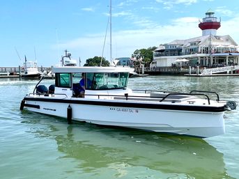 White pilothouse powerboat gliding through calm green harbor water past a waterfront marina and a red-and-white lighthouse on a sunny day.