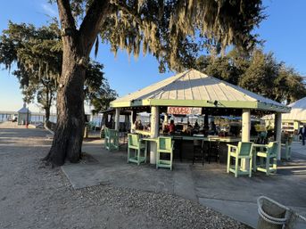 Outdoor waterfront pavilion bar with pastel-green high chairs around a central counter under a metal roof, large oak draped in Spanish moss, and marina docks and boats in the background at golden hour.