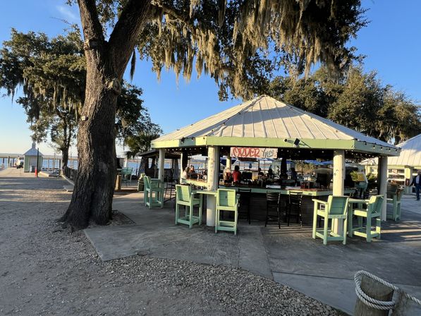 Outdoor waterfront pavilion bar with pastel-green high chairs around a central counter under a metal roof, large oak draped in Spanish moss, and marina docks and boats in the background at golden hour.