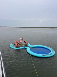Four people relaxing and waving on a bright blue inflatable floating island in calm coastal bay waters, tethered to a boat under an overcast sky.