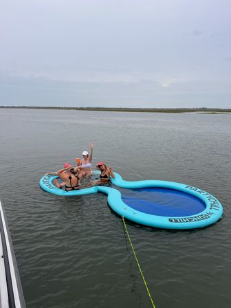 Four people relaxing and waving on a bright blue inflatable floating island in calm coastal bay waters, tethered to a boat under an overcast sky.