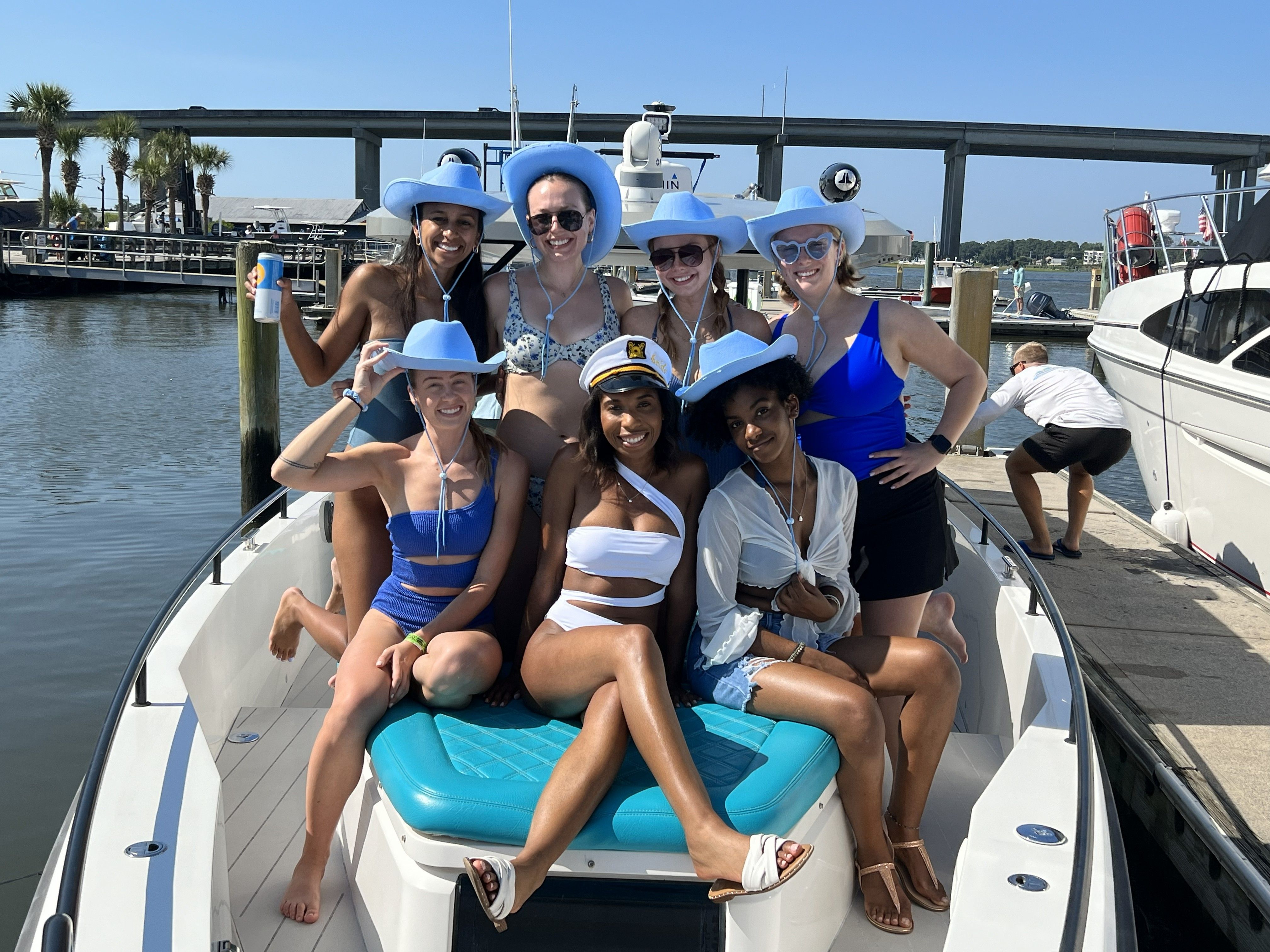 Cheerful group of seven women in swimwear and matching light-blue cowboy hats, one wearing a captain’s hat, posing on the bow of a boat at a sunny coastal marina with a bridge and dock in the background.