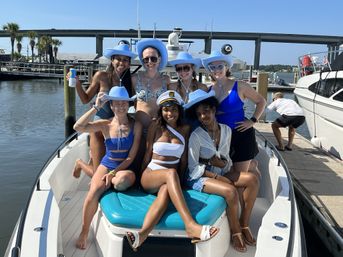 Cheerful group of seven women in swimwear and matching light-blue cowboy hats, one wearing a captain’s hat, posing on the bow of a boat at a sunny coastal marina with a bridge and dock in the background.