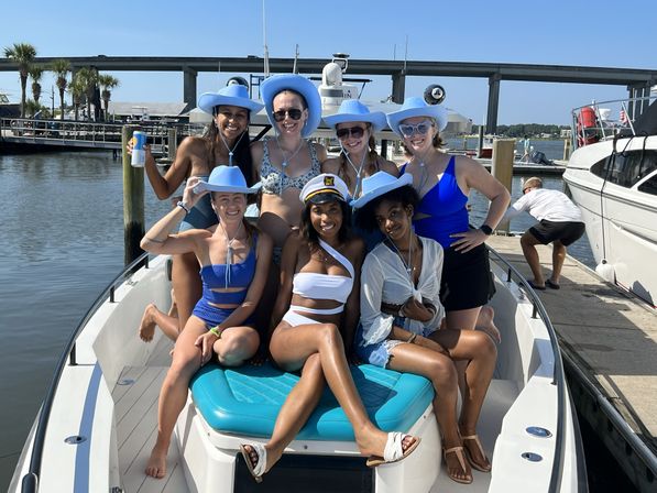 Cheerful group of seven women in swimwear and matching light-blue cowboy hats, one wearing a captain’s hat, posing on the bow of a boat at a sunny coastal marina with a bridge and dock in the background.