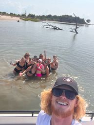 Boat selfie of a red-haired man in sunglasses and cap with a group of friends in bikinis waist-deep in a calm coastal inlet near a sandy shore and driftwood