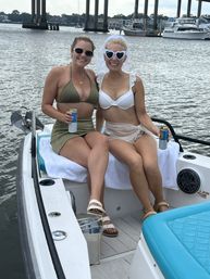 Two friends in bikinis smiling on the bow of a small boat at a marina beneath a concrete bridge, wearing sunglasses and holding canned drinks — playful bachelorette boat outing