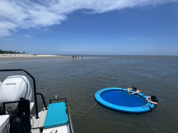 Motorboat anchored by a shallow coastal sandbar with people walking on the beach and two people relaxing in a round blue inflatable floating pool under a clear blue sky.