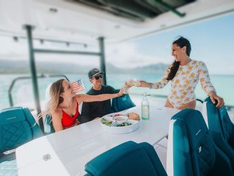 Three friends toasting drinks on a sunny boat deck with teal seats, a snack platter, small American flag, and turquoise ocean and mountains in the background.