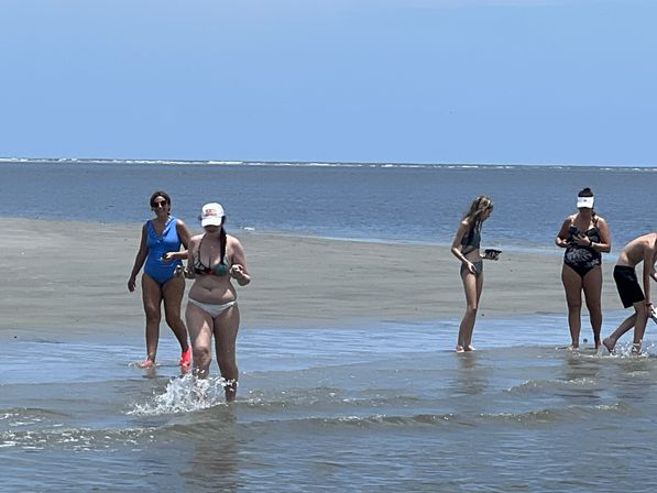 Group of beachgoers in swimsuits wading and splashing in shallow ocean water along a wide sandy shoreline on a sunny summer day