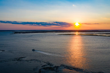 Aerial view of a small motorboat crossing a calm coastal estuary at sunset, golden sun reflecting on water with marsh islands and distant horizon.