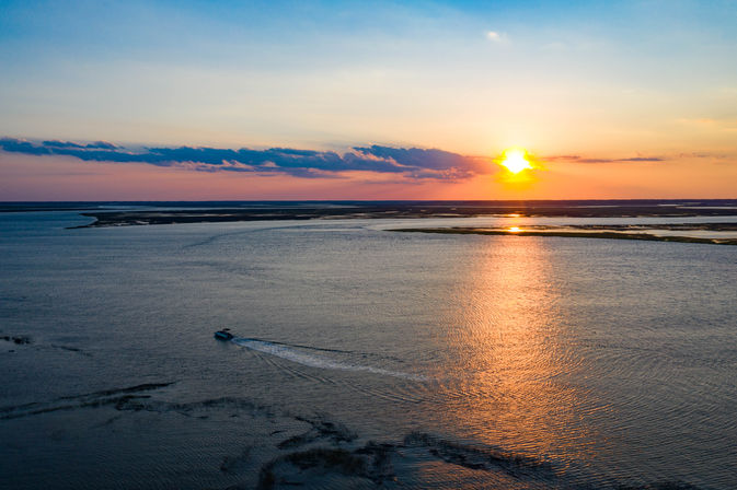Aerial view of a small motorboat crossing a calm coastal estuary at sunset, golden sun reflecting on water with marsh islands and distant horizon.